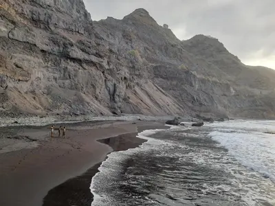 two people walking on a beach next to a mountain at Sinagoga Guesthouse in Ribeira Grande